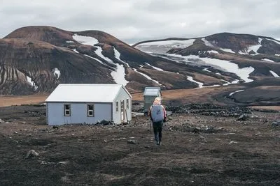 A hiker walking towards a small mountain hut in a dark volcanic landscape with snow patches