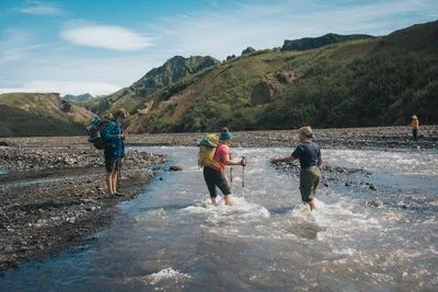 Hikers with trekking poles wading through a shallow glacial river in a rocky valley